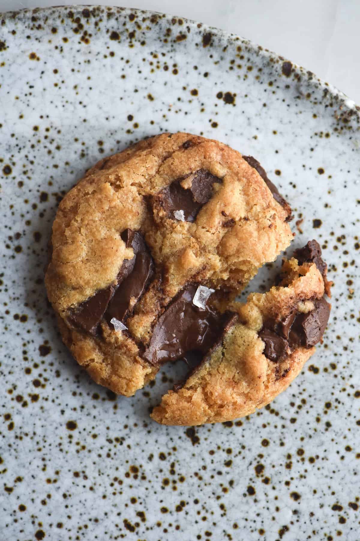 An aerial view of a gluten free vegan choc chip cookie on a white speckled ceramic plate. The cookie has been snapped down the middle, exposing the melted chocolate that lies within. It is topped with a sprinkle of sea salt flakes