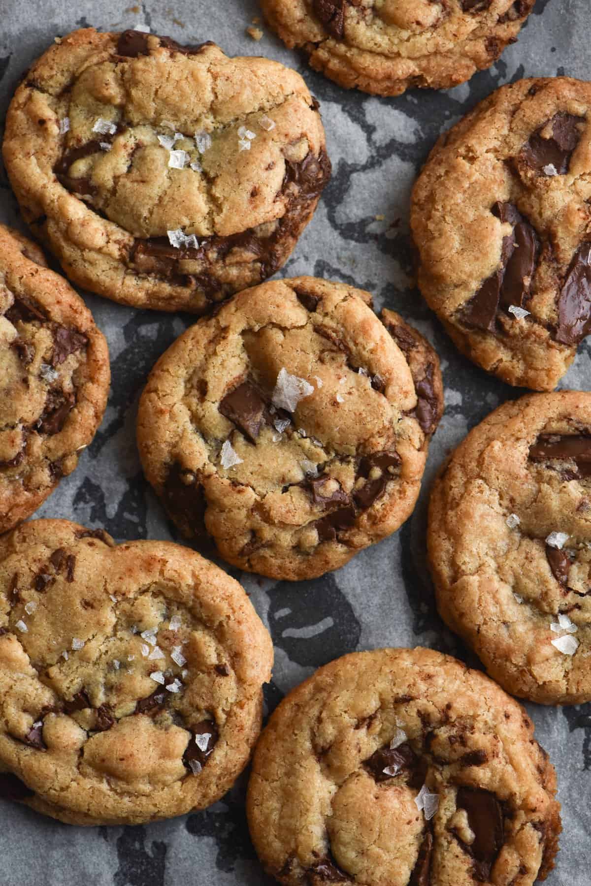 A close up shot of gluten-free vegan choc-chip cookies topped with flaky salt. They sit atop a black baking tray with baking paper on top.