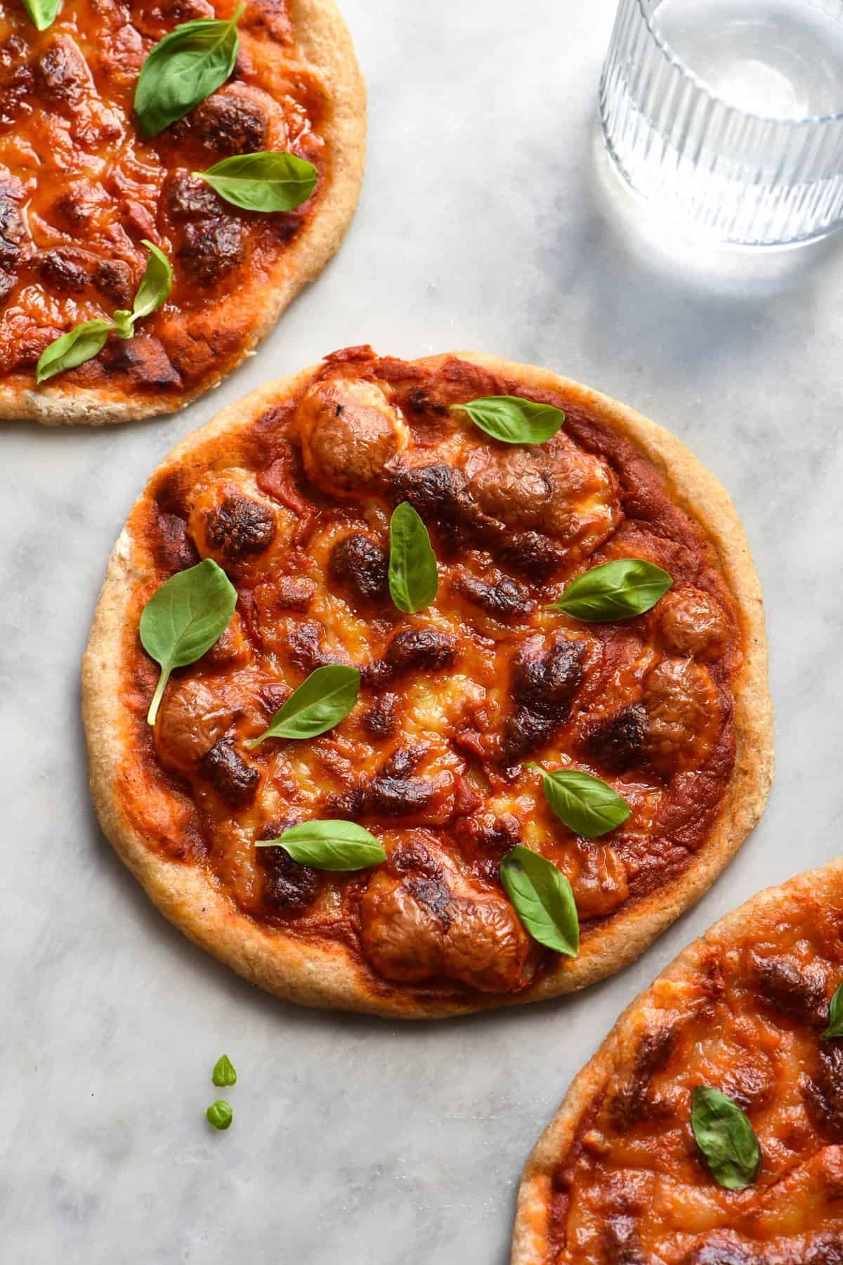 An aerial view of three gluten free sourdough pizza bases topped with FODMAP friendly pizza sauce and melty cheese. The pizzas sit in a diagonal pattern on the table and a glass of water sits in the top right hand side of the image