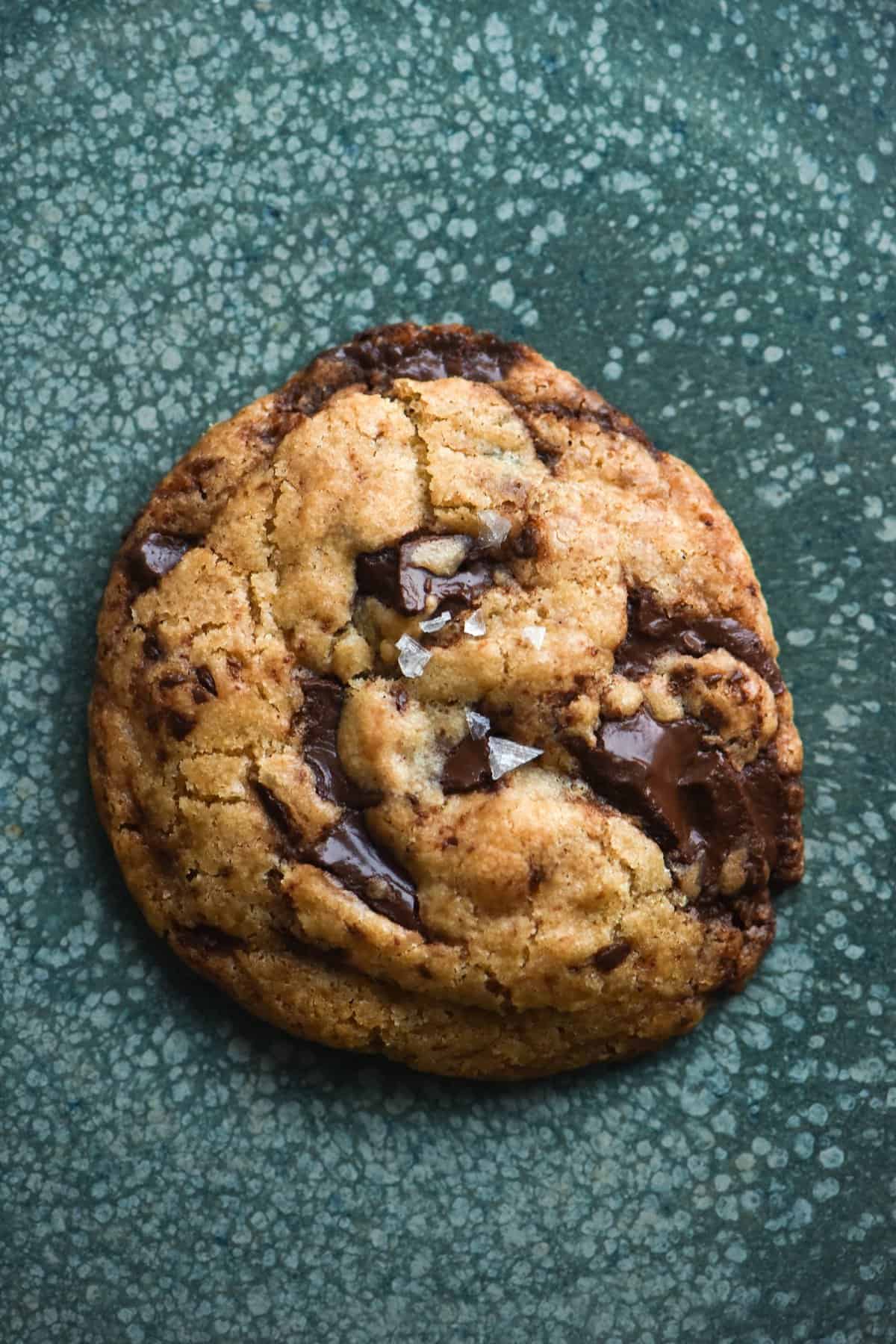 An aerial view of a gluten free vegan choc chip cookie atop a turquoise green ceramic speckled plate. The cookie is studded with melted dark chocolate and topped with sea salt flakes