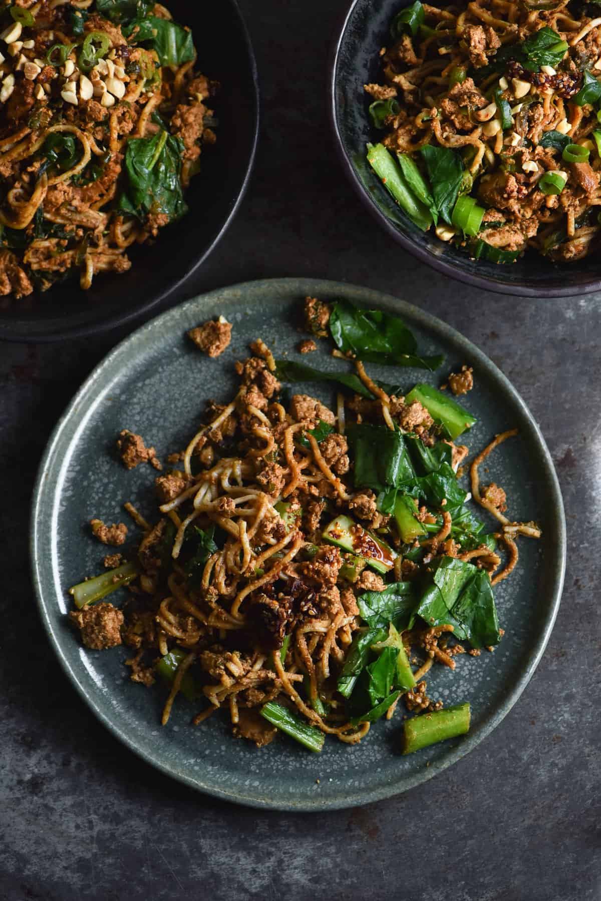 An aerial view of three plates of FODMAP friendly vegetarian Dan Dan style noodles. The noodles sit against a blue mottled backdrop