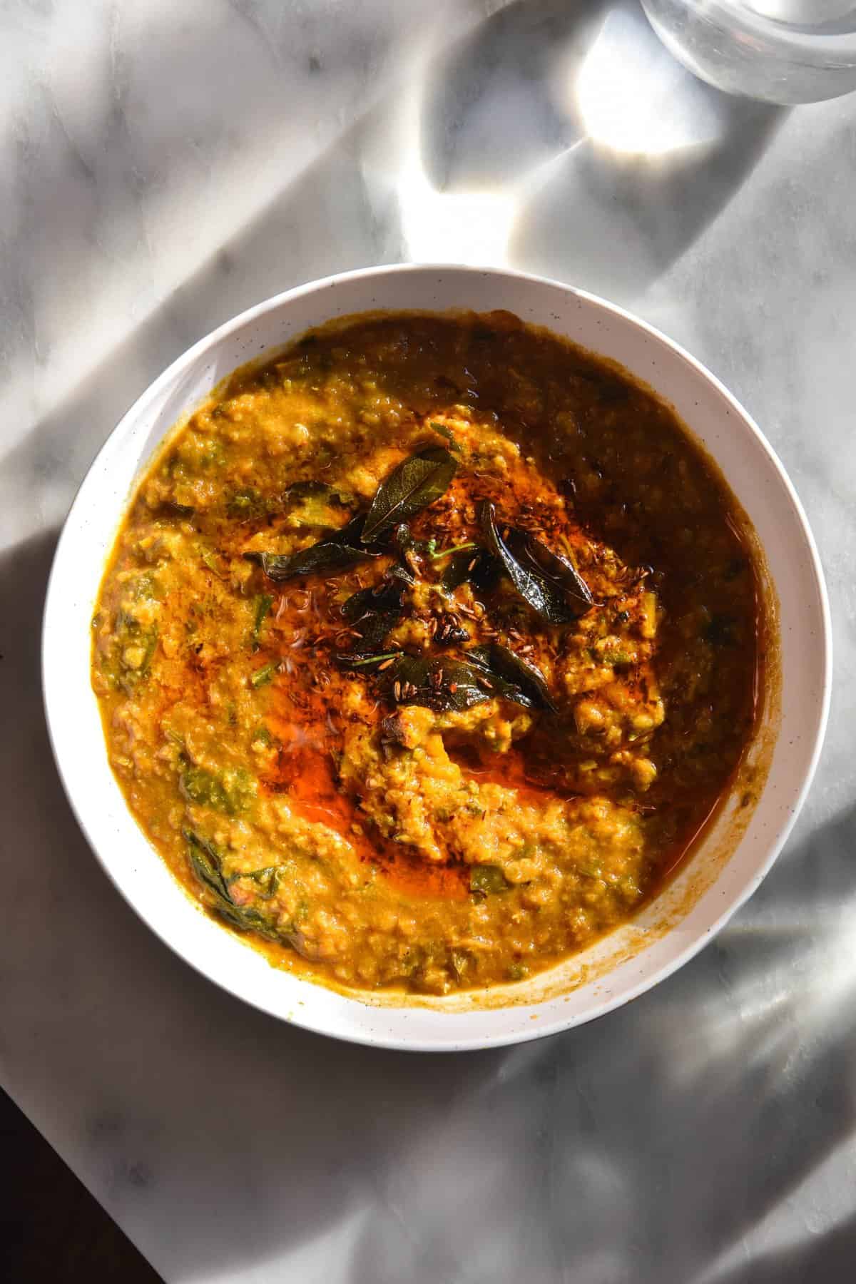 A close up aerial view of a white bowl filled with FODMAP friendly daal. The daal is topped with fried curry leaves, a chilli tadka and chopped coriander. The dish sits against a white marble table.