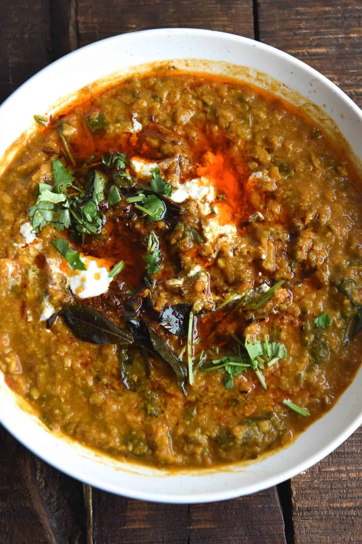 A close up aerial view of a white bowl filled with FODMAP friendly daal. The daal is topped with fried curry leaves, a chilli tadka, chopped coriander and a little bit of yoghurt. The dish sits against a dark wood table.