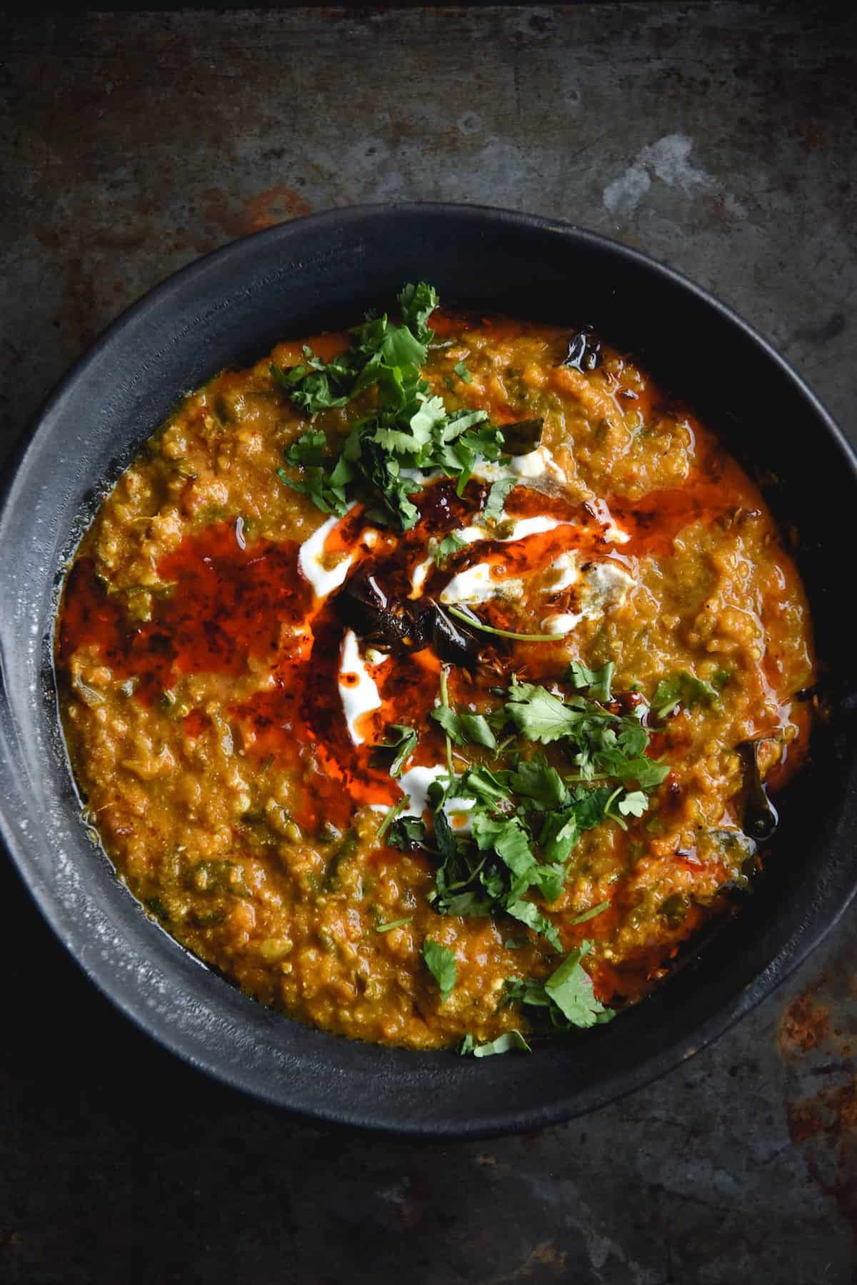 A close up aerial view of a dark ceramic bowl filled with FODMAP friendly dahl. The dahl is topped with a chilli oil tadka, fried curry leaves, chopped coriander and a bit of yoghurt. The bowl is to the right of the image and sits on a dark blue backdrop