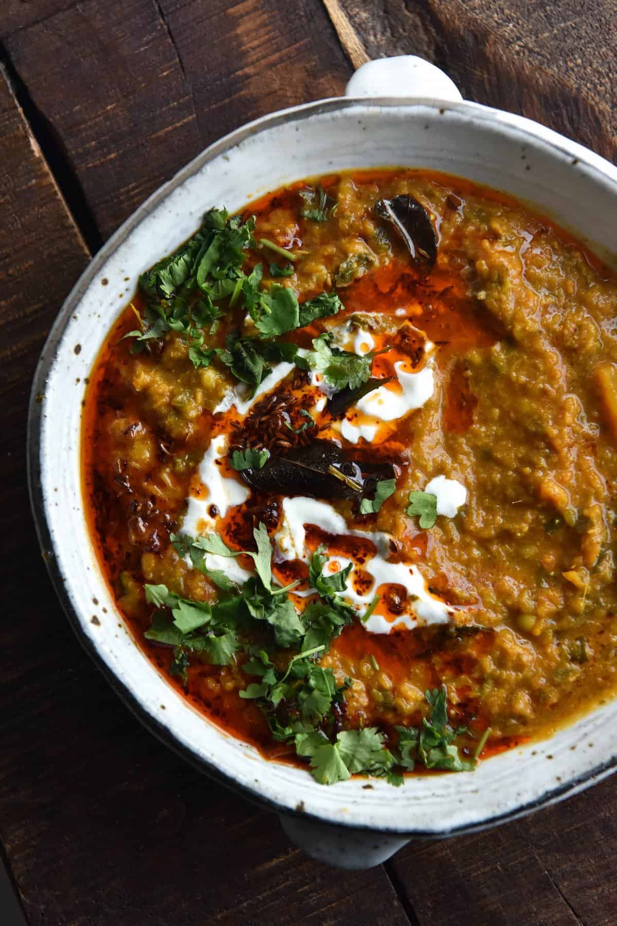 An aerial view of a white speckled ceramic bowl of FODMAP friendly daal. The daal is topped with a chilli oil tadka, fried curry leaves, coriander and some yoghurt. The bowl sits to the right of the image and sits against a dark wooden textured backdrop