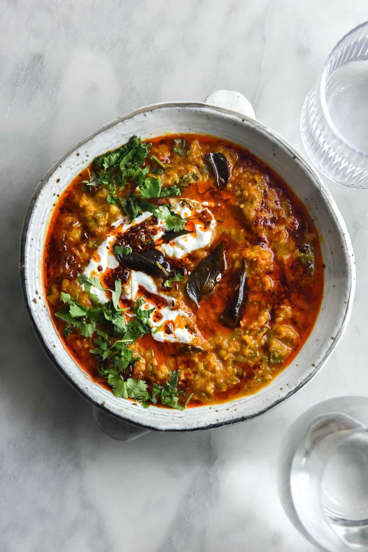 An aerial view of a white speckled ceramic bowl of FODMAP friendly daal. The daal is topped with a chilli oil tadka, fried curry leaves, coriander and some yoghurt. The bowl sits in the centre of a white marble table and two sunlit water glasses sit to the edge of the image