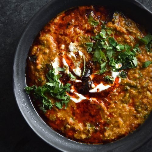 A close up aerial view of a dark ceramic bowl filled with FODMAP friendly dahl. The dahl is topped with a chilli oil tadka, fried curry leaves, chopped coriander and a bit of yoghurt. The bowl is to the right of the image and sits on a dark blue backdrop