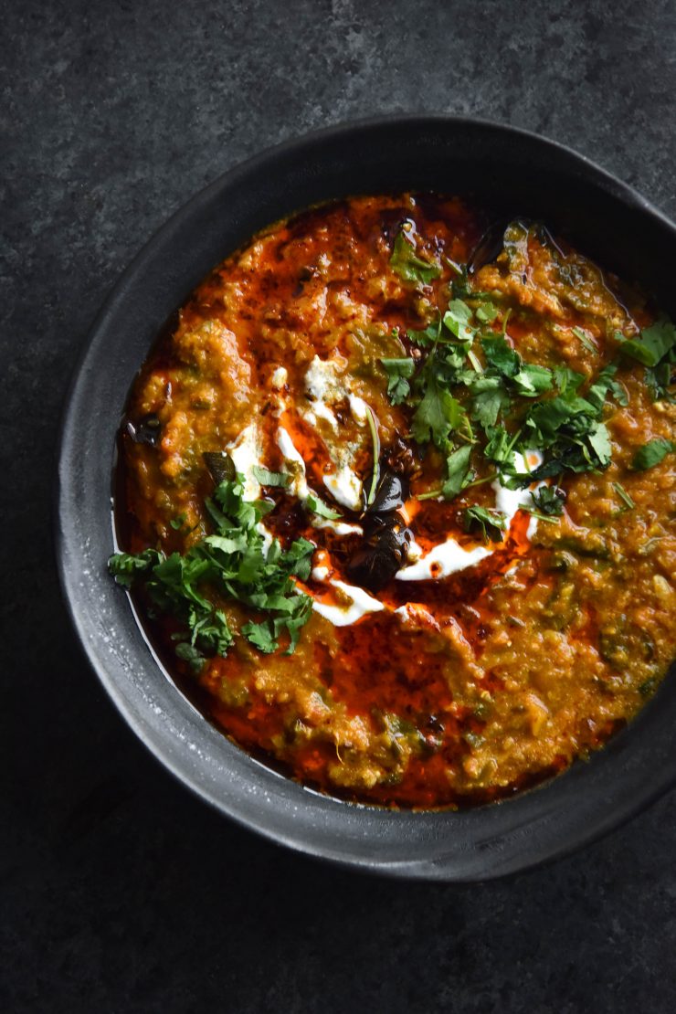 A close up aerial view of a dark ceramic bowl filled with FODMAP friendly dahl. The dahl is topped with a chilli oil tadka, fried curry leaves, chopped coriander and a bit of yoghurt. The bowl is to the right of the image and sits on a dark blue backdrop