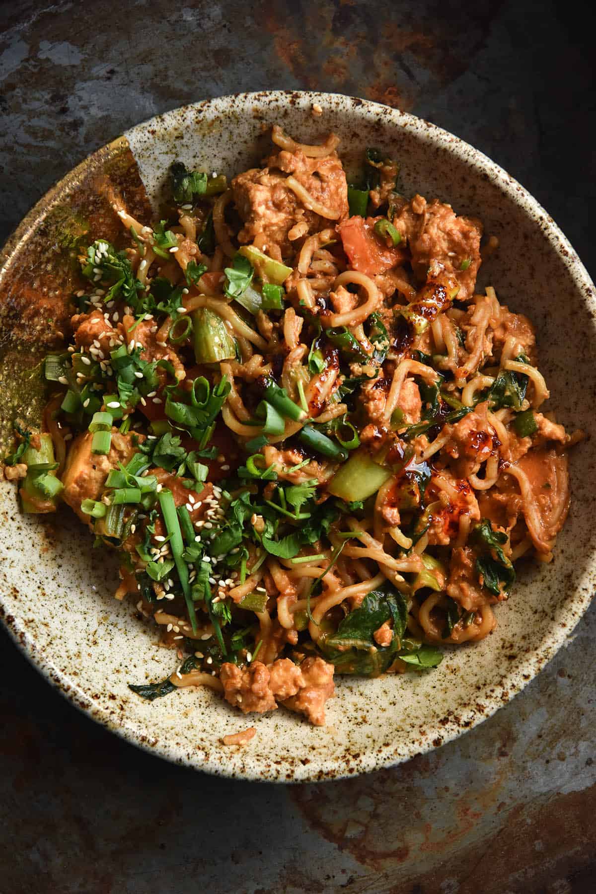 A close up aerial view of a speckled ceramic bowl filled with vegetarian peanut butter noodles. The noodles are topped with toasted sesame seeds, spring onion and coriander and sit against a mottled grey backdrop