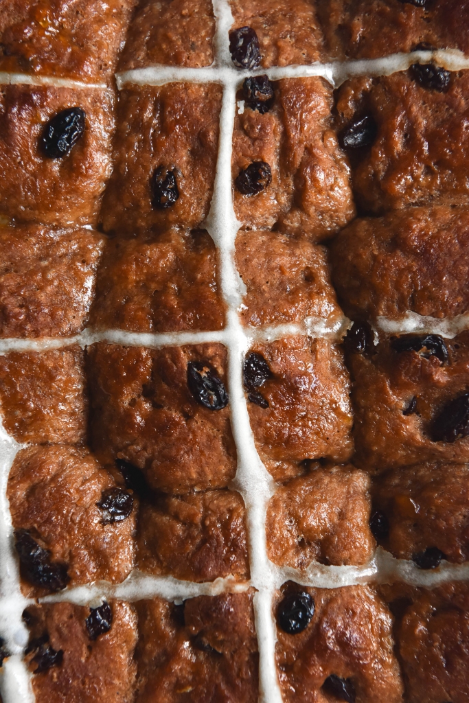 A close up aerial view of 9 gluten free vegan hot cross buns sitting tightly in a tray. They are deeply golden and speckled with raisins