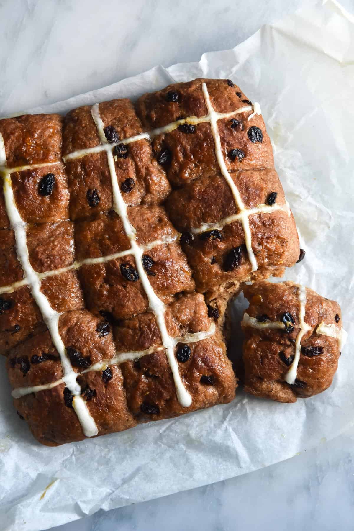 An aerial view of a sheet of gluten free, FODMAP friendly hot cross buns. The buns sit atop a white marble table on a crinkled sheet of baking paper