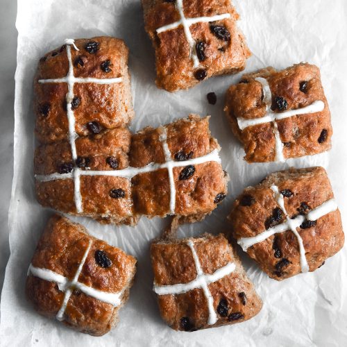 An aerial view of 9 gluten free vegan hot cross buns. The buns are strewn across a light piece of baking paper on a white marble table