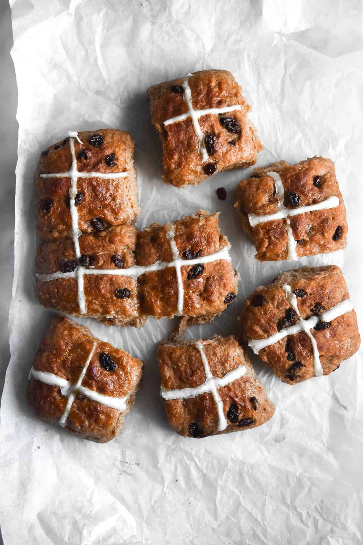 An aerial view of 9 gluten free vegan hot cross buns. The buns are strewn across a light piece of baking paper on a white marble table