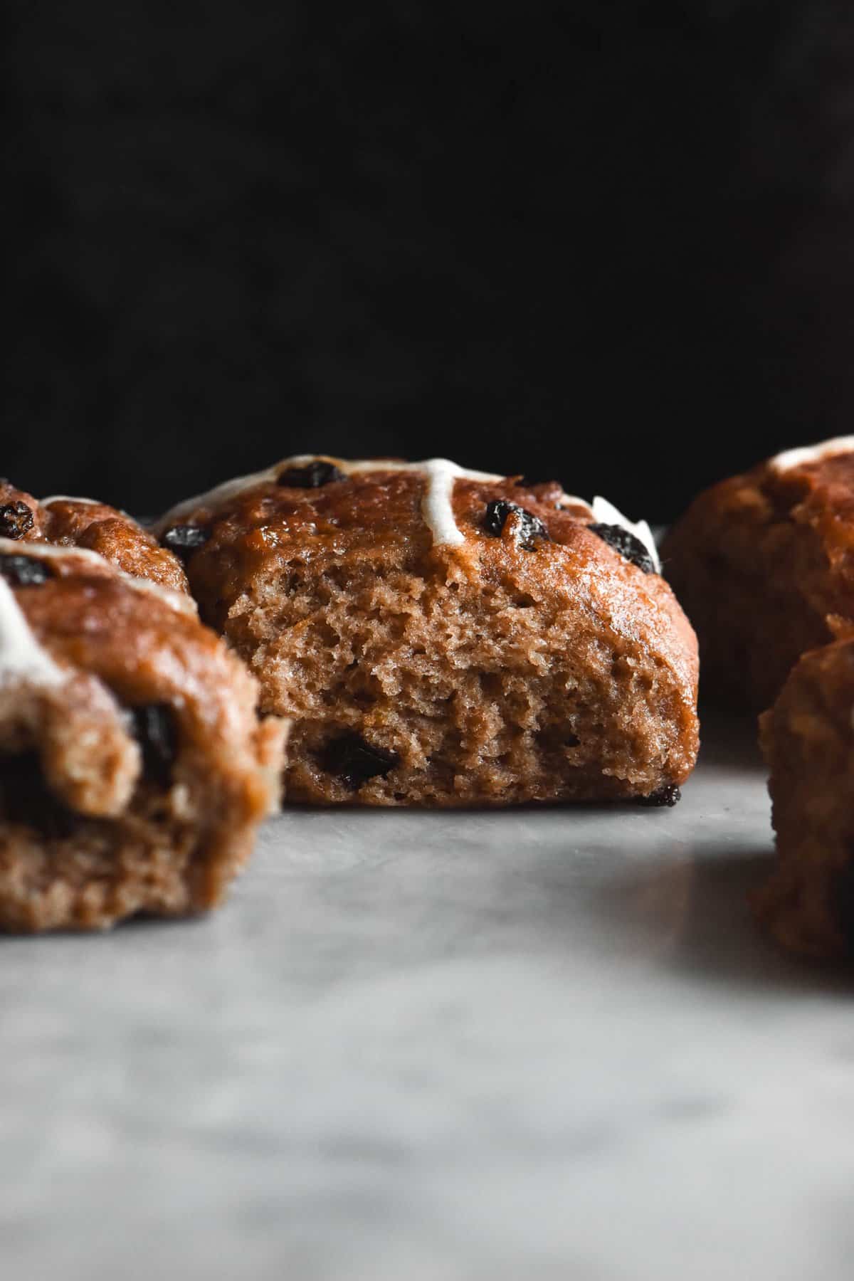 A side on view of gluten free vegan hot cross buns. The buns are deeply spiced and flecked with raisins. They sit atop a white marble table, contrasted against a black backdrop