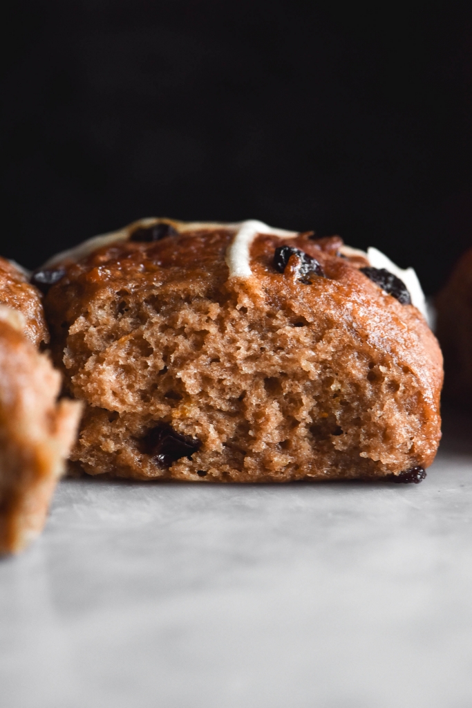 A side on view of a gluten free hot cross bun. The bun sits atop a white marble table against a black backdrop. A raisin peeks out from the soft, fluffy spice dough.