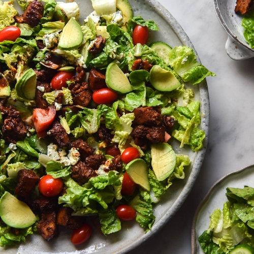 An aerial view of a vegetarian Cobb salad topped with sweet and smoky tofu. A big plate of salad sits to the left of the image, and two smaller bowls sit to the right