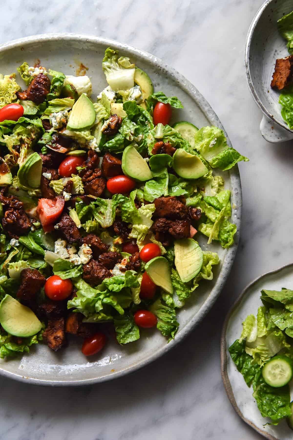 An aerial view of a vegetarian Cobb salad topped with sweet and smoky tofu. A big plate of salad sits to the left of the image, and two smaller bowls sit to the right