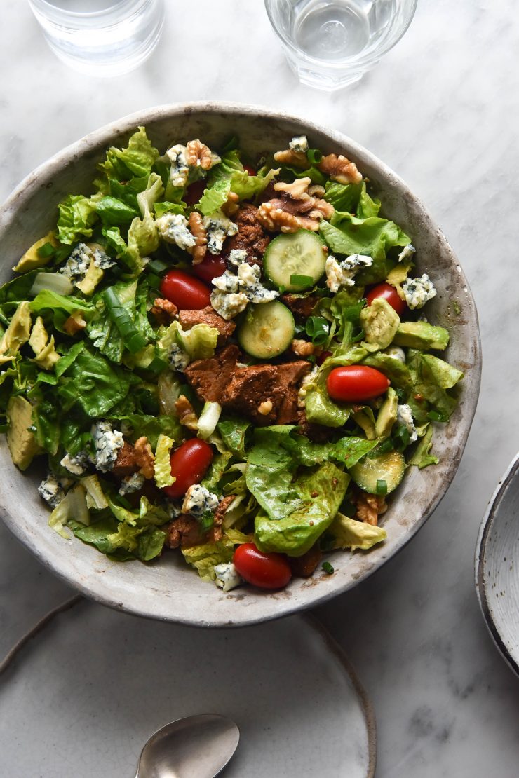 An aerial view of a vegetarian Cobb style salad in a light grey ceramic bowl. The salad sits atop a white marble table, with white ceramics strewn around the table. Two glasses of water sit at the top of the image