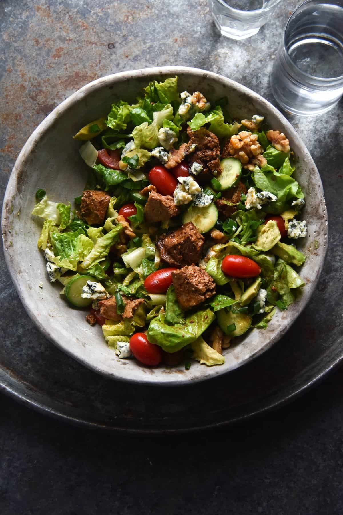 An aerial view of a vegetarian Cobb style salad in a light grey ceramic bowl. The salad sits atop a mottled blue tray on a navy coloured backdrop. Two glasses of water sit to the right hand side of the image