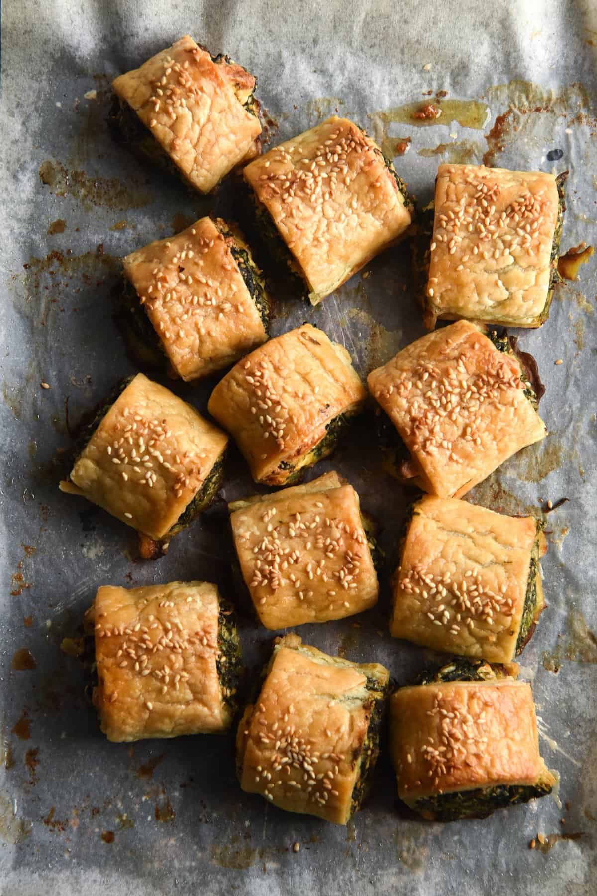 An aerial view of a tray of gluten free spinach and feta rolls. The rolls are casually strewn in a bundle on a baking paper lined baking tray. They are golden brown and topped with toasted sesame seeds