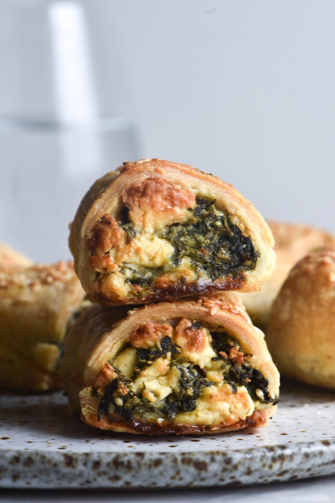A side on macro photo of a stack of two gluten free spinach and feta sausage rolls. The rolls sit atop a white speckled ceramic plate, surrounded by other rolls. Golden feta and pine nuts poke out of the filling, and the top of the pastry is sprinkled with toasted sesame seeds. A glass of water sits against the white backdrop in the background.