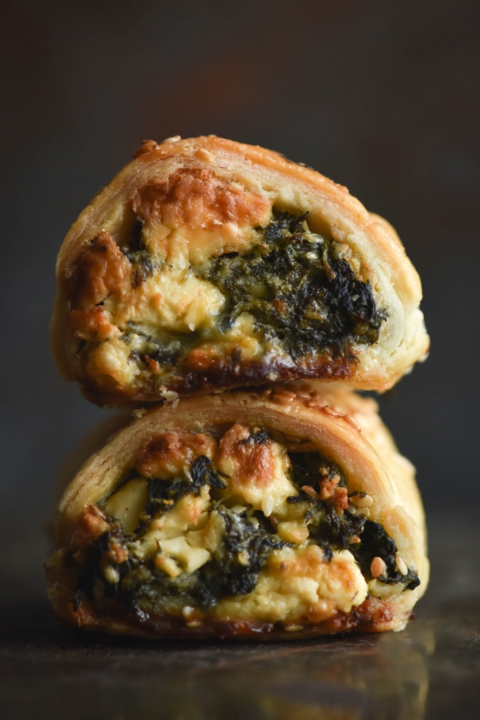 A side on macro photo of a stack of two gluten free spinach and feta sausage rolls. The rolls sit atop a dark mottled backdrop and in front of a mottled grey and rusting background. Golden feta and pine nuts poke out of the filling, and the top of the pastry is sprinkled with toasted sesame seeds.