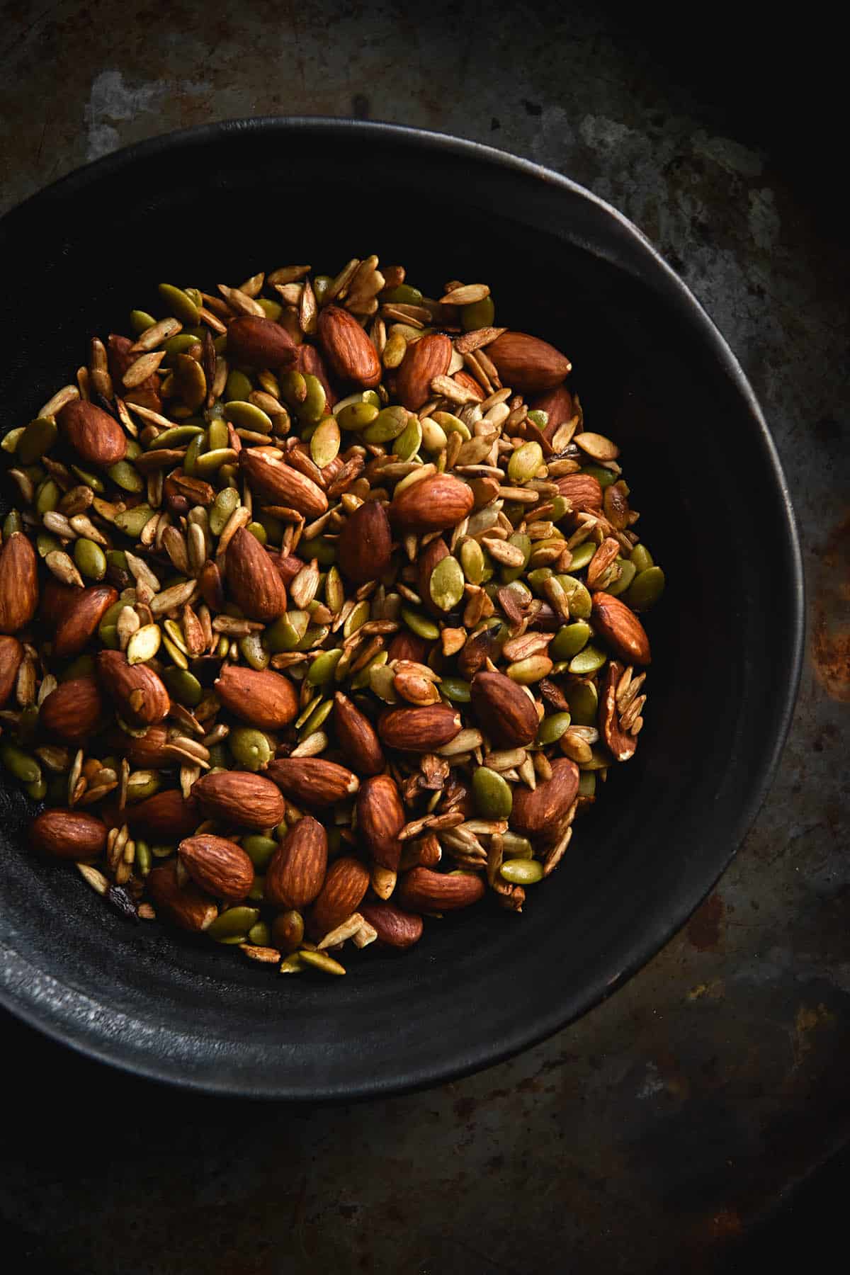 a close up aerial shot of a dark ceramic bowl filled with tamari nut mix. The nut mix consists of almonds, pepitas and sunflower seeds, and they are all mixed together peeking out of the open faced bowl. The bowl sits atop a mottled grey background and the image has a warm but moody feeling