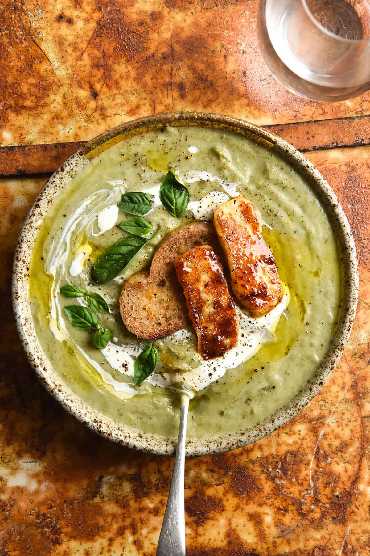 An aerial view of a speckled ceramic bowl of FODMAP friendly leek and potato soup. The soup is a light green because it uses leek greens to keep it FODMAP friendly. It is topped with a swirl of cream, truffle oil, a gluten free crouton, grilled haloumi and basil leaves. It sits atop a mottled bright rusty backdrop and a glass of water sits in the top right hand corner