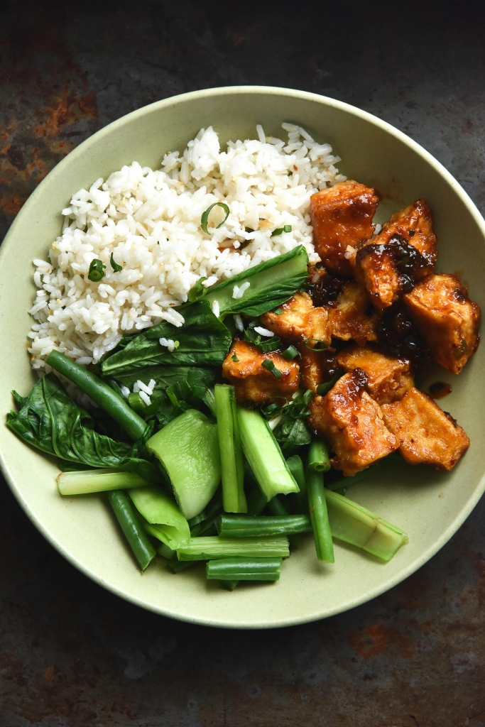 A close up aerial view of a bowl of sweet and sticky ginger tofu with ginger sesame rice and Asian greens. The meal sits in a pale green open faced bowl against a mottled navy coloured backdrop