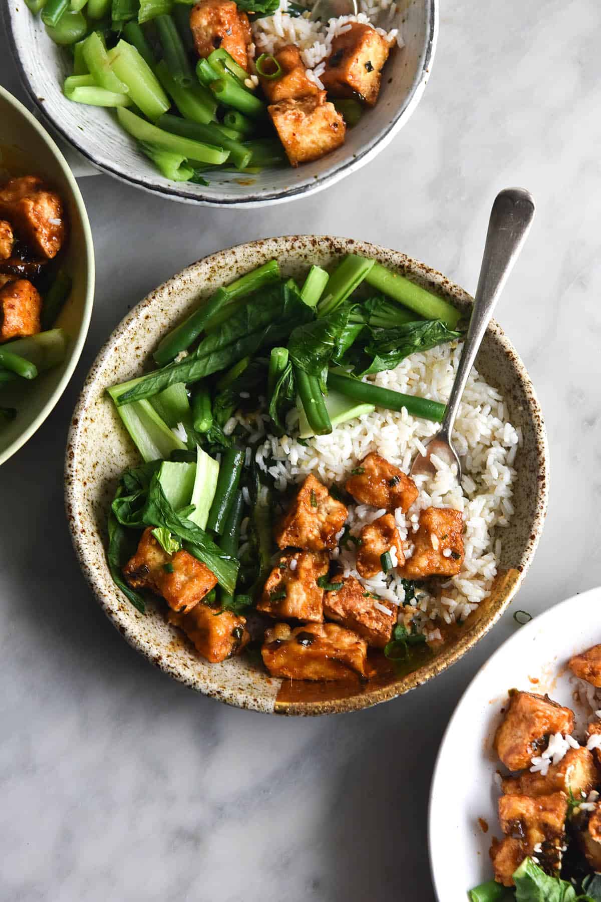 A close up aerial view of four bowls of sticky ginger tofu, Asian greens and sesame ginger rice sitting atop a white marble table in a casual fashion. The central bowl is a ceramic beige speckled one, and has a fork poking out of the top left corner of the bowl