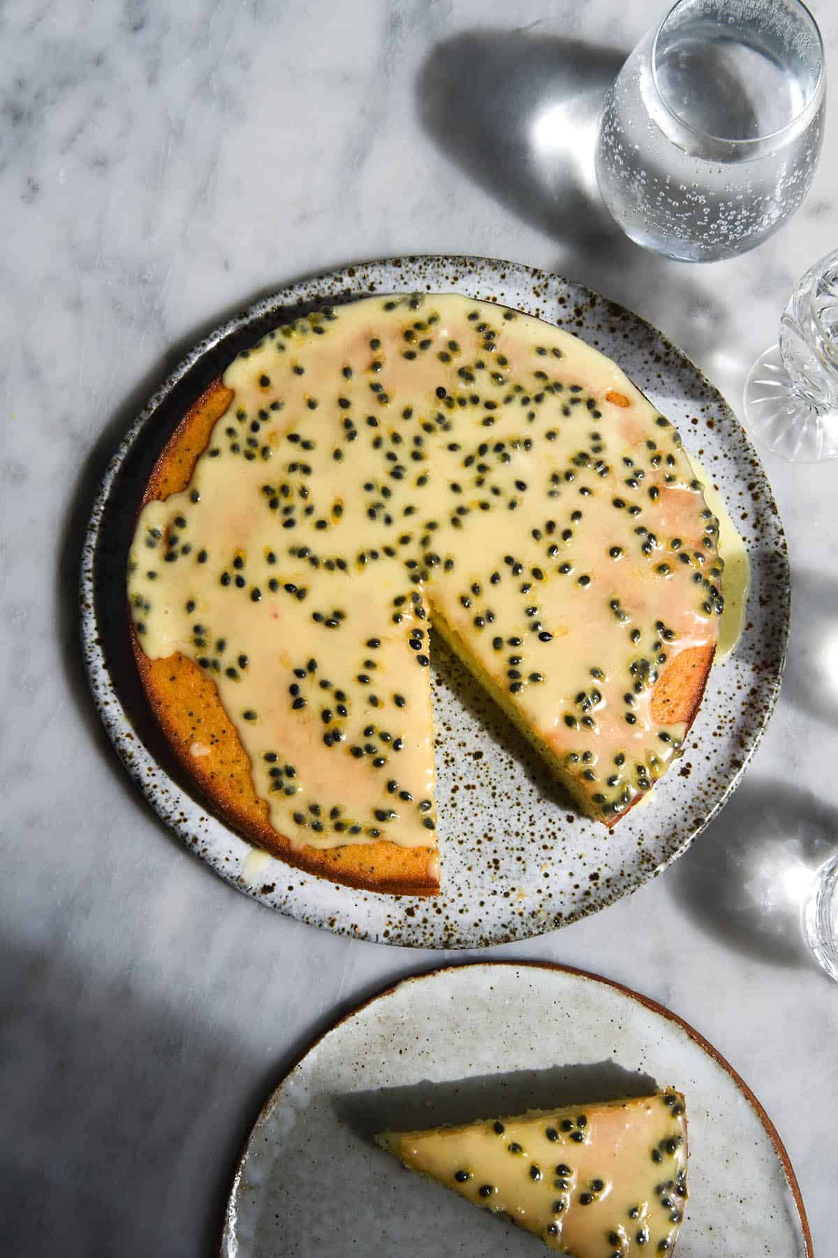 An aerial view of an easy gluten free vanilla cake topped with passionfruit icing. The cake sits atop a white marble table in harsh sunlight and is surrounded by glasses of water and a white ceramic plate with a slice of cake
