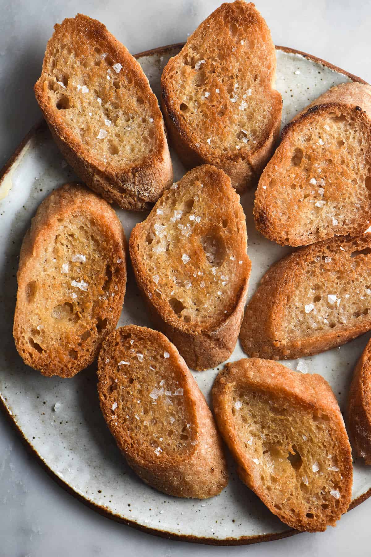 An aerial view of slices of gluten free baguette that have been fried in garlic infused ghee to create a FODMAP friendly garlic bread. The slices are arranged on a white ceramic plate and sprinkled with flaky sea salt.