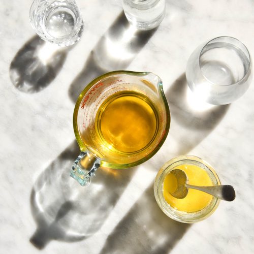 An aerial view of two jugs of freshly made garlic infused ghee sit on a white marble table in harsh sunlight. They are surrounded by water glasses and the light shines through creating shadows on the marble