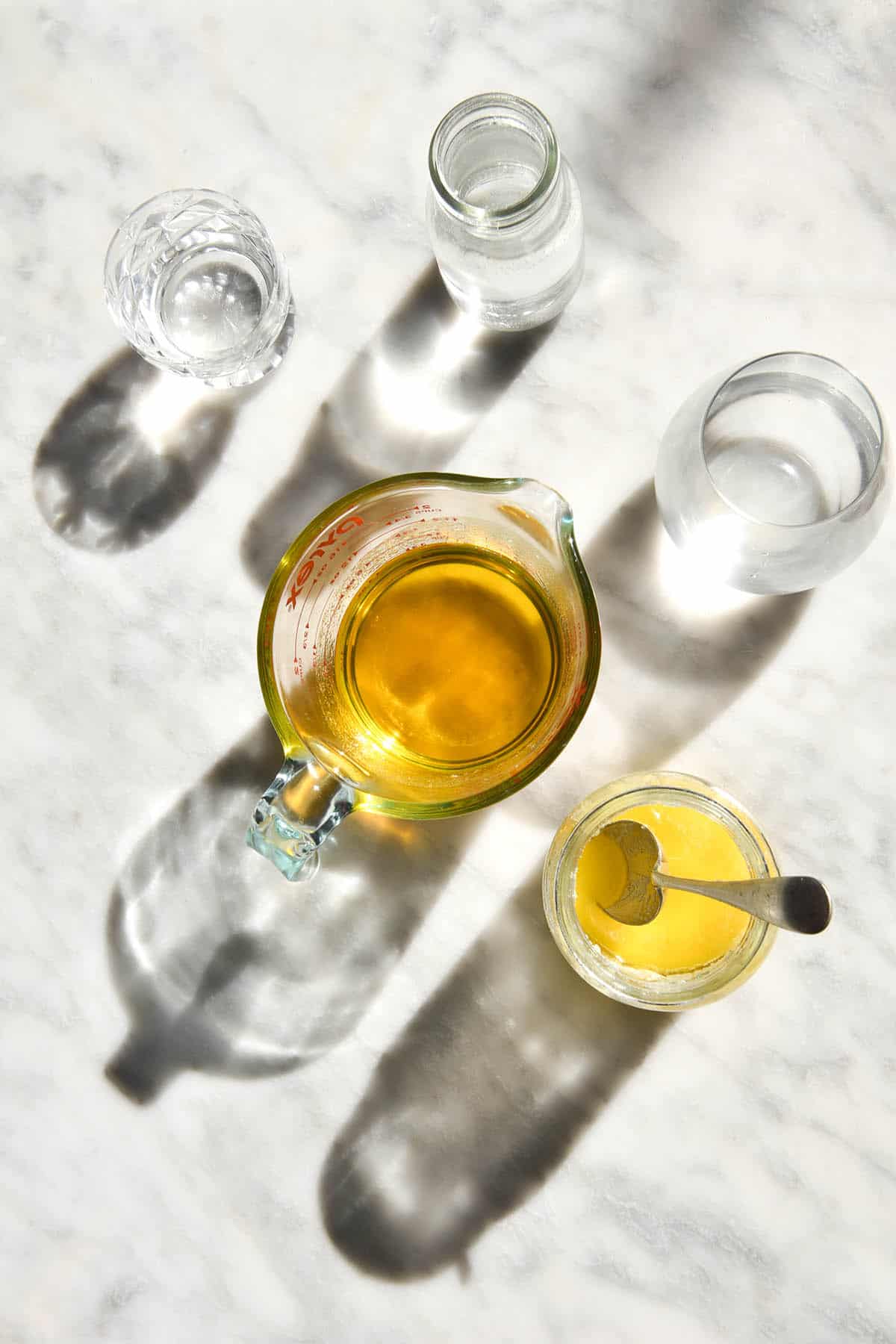 An aerial view of two jugs of freshly made garlic infused ghee sit on a white marble table in harsh sunlight. They are surrounded by water glasses and the light shines through creating shadows on the marble