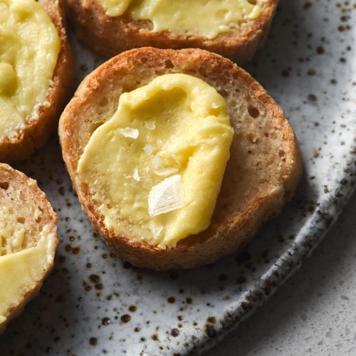 A close up, side on view of a plate of sliced baguette rounds, each topped with a slathering of whipped garlic infused ghee and a sprinkle of flaky sea salt. The baguette rounds sit atop a white speckled ceramic plate on a white background.