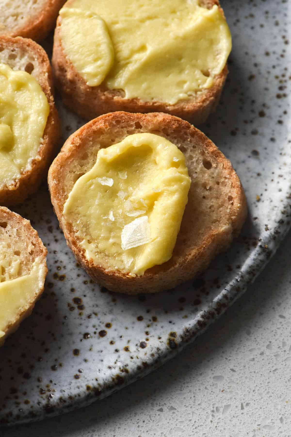 A close up, side on view of a plate of sliced baguette rounds, each topped with a slathering of whipped garlic infused ghee and a sprinkle of flaky sea salt. The baguette rounds sit atop a white speckled ceramic plate on a white background.