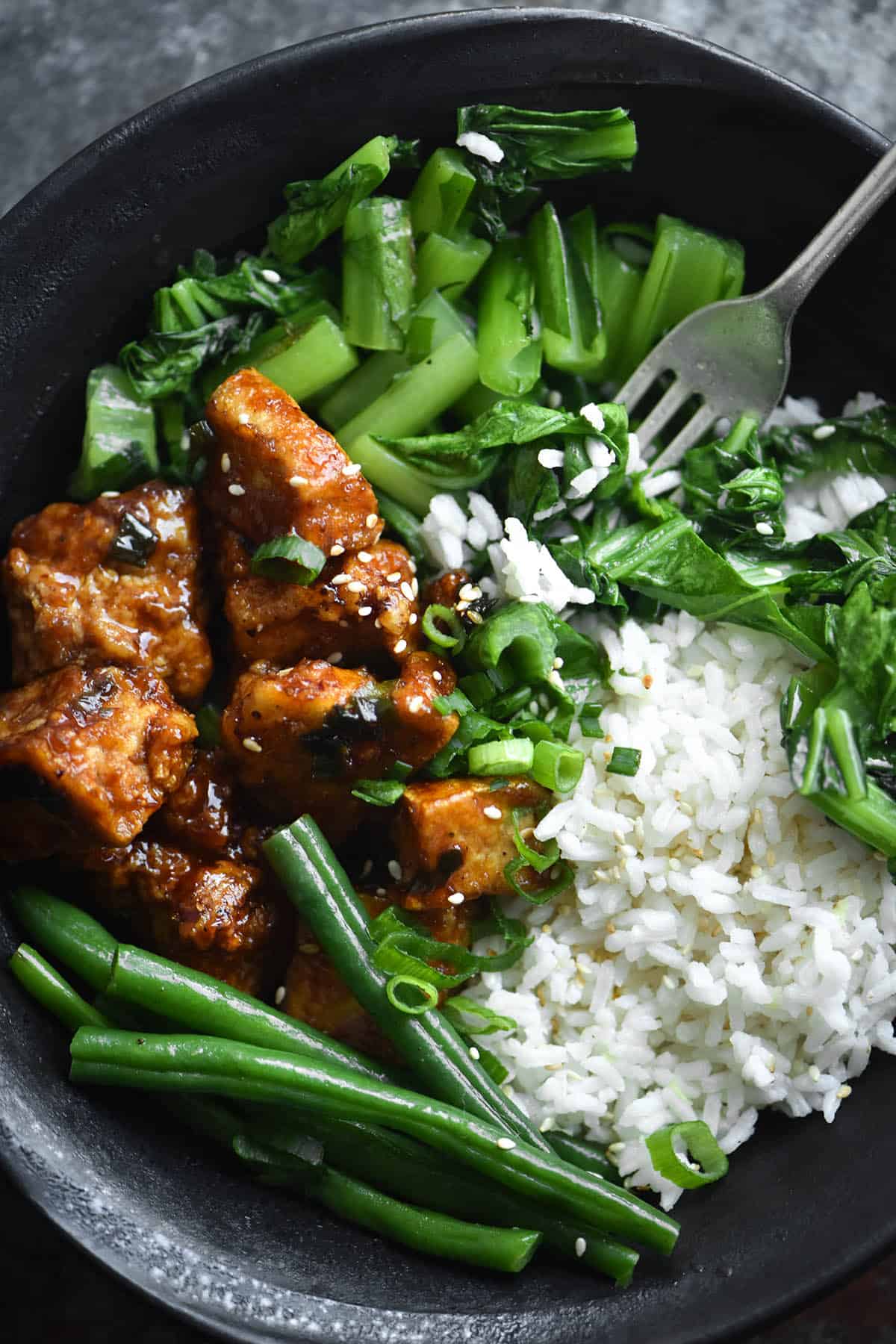 A close up aerial view of a bowl of sweet and sticky ginger tofu with ginger sesame rice and Asian greens. The meal sits in a dark blue ceramic bowl against a mottled navy coloured backdrop