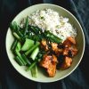 A close up aerial view of a bowl of sweet and sticky ginger tofu with ginger sesame rice and Asian greens. The meal sits in a pale green open faced bowl against a textured olive linen backdrop