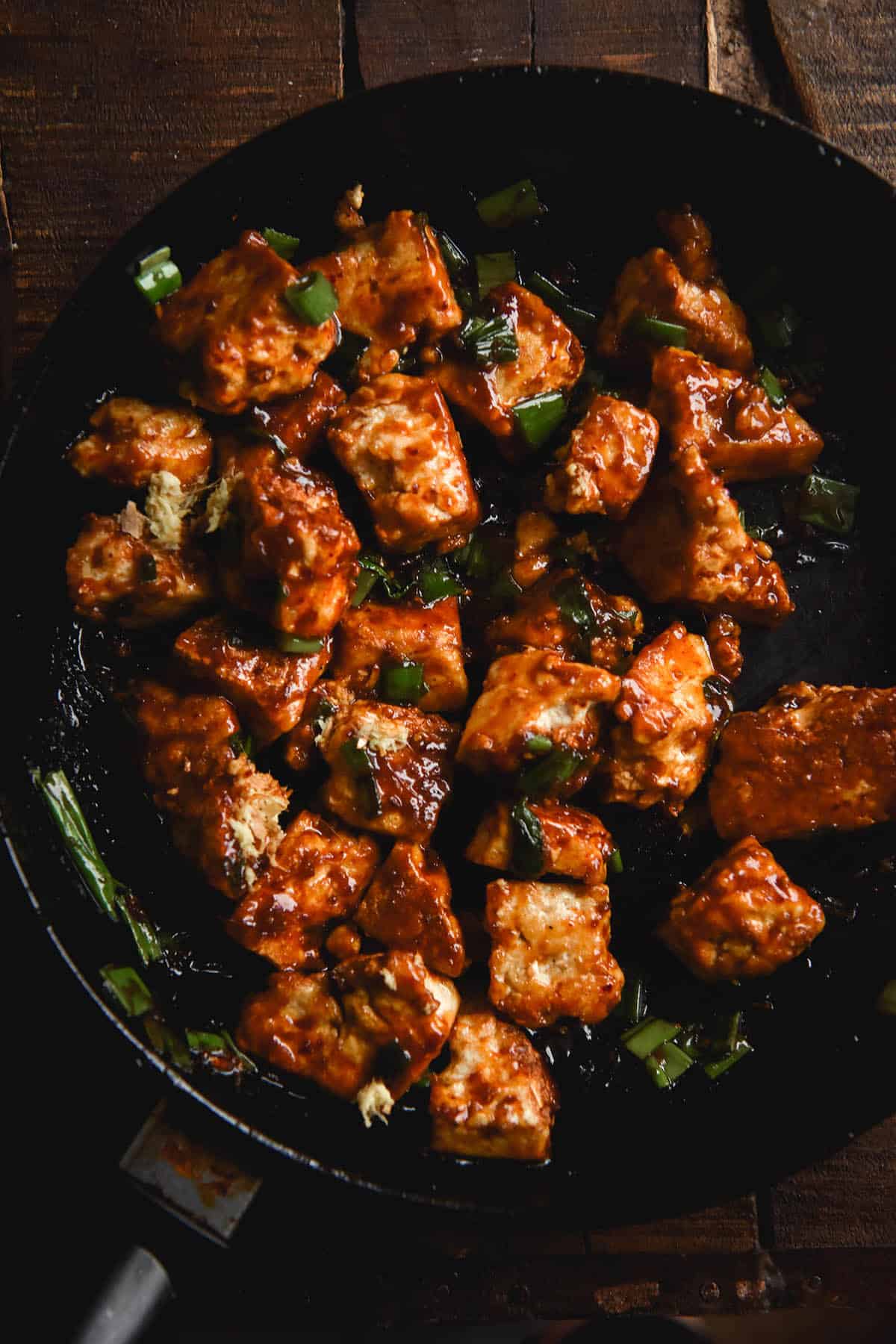 A close up aerial view of bite sized chunks of sticky ginger glazed tofu in a pan against a dark wooden backdrop. The tofu is a deep red/orange colour and is specked with spring onion greens.