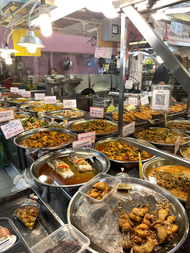 A food stall at Or Tor Kor market in Bangkok. There are lots of different large round silver trays upon which various dishes sit, mainly fish ones.