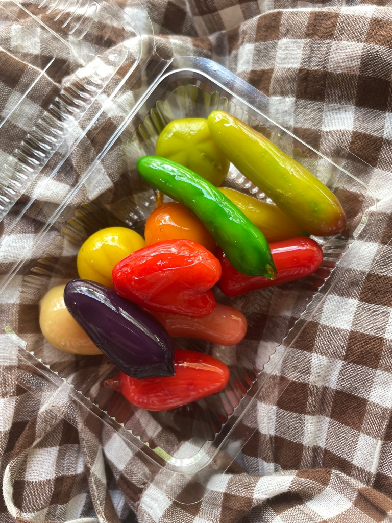 A plastic container of kanom luk chup, a colourful Thai dessert made with mung beans and sugar. The dessert is crafted into miniature vegetables of all different colours - green chilli, tomato and eggplant, to name a few. The container sits on the lap of a woman wearing a light brown and white checked dress.