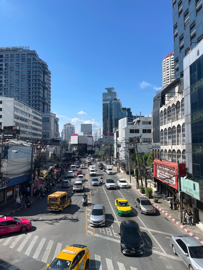 A view of the streetscape from the BTS skyline walking platform in Bangkok. Cars are in motion across a pedestrian crossing. Skyscrapers and a blue sky form the backdrop