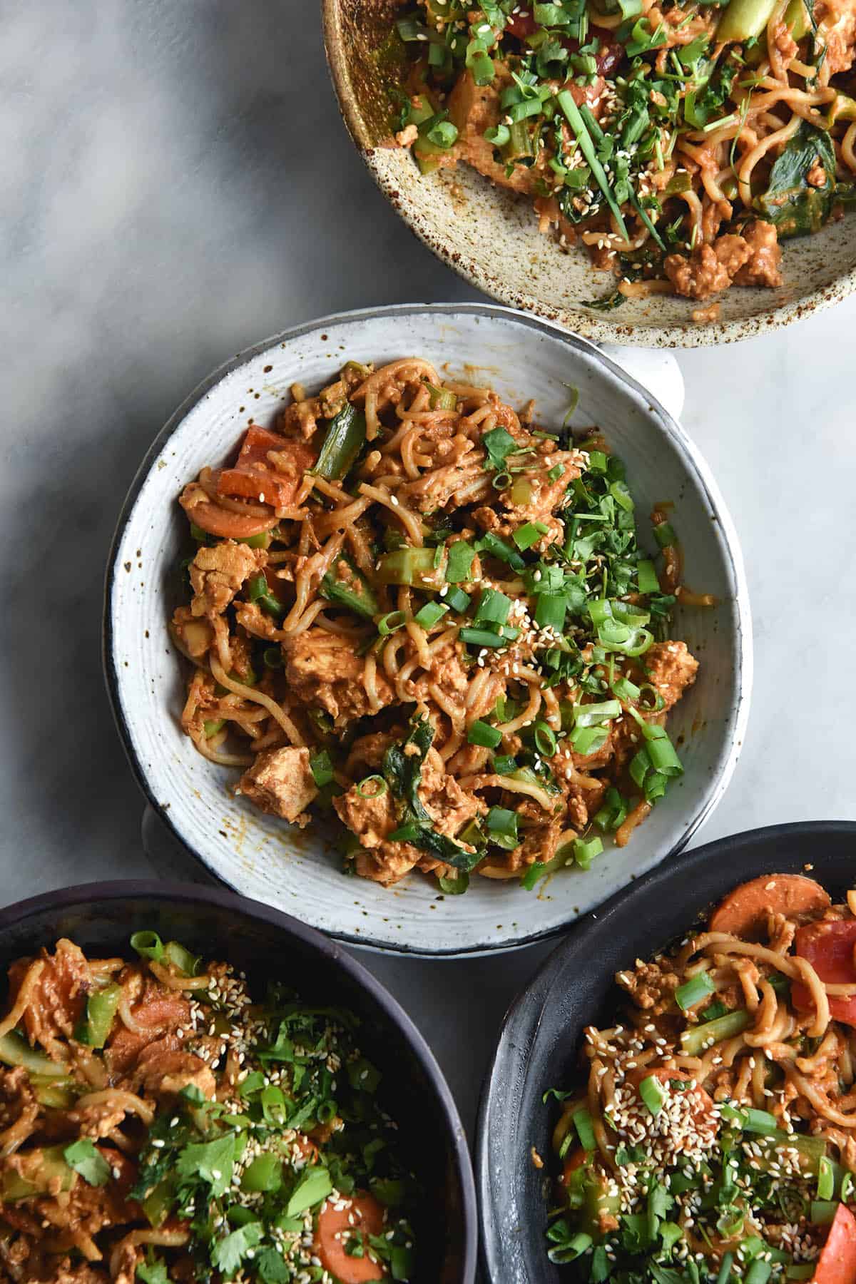 An aerial view of four ceramic bowls filled with FODMAP friendly peanut butter stir fry. The stir fry is filled with chunks of tofu and vegetables and topped with sesame seeds and spring onions. The bowls are all different colours and shapes, sitting atop a white marble backdrop