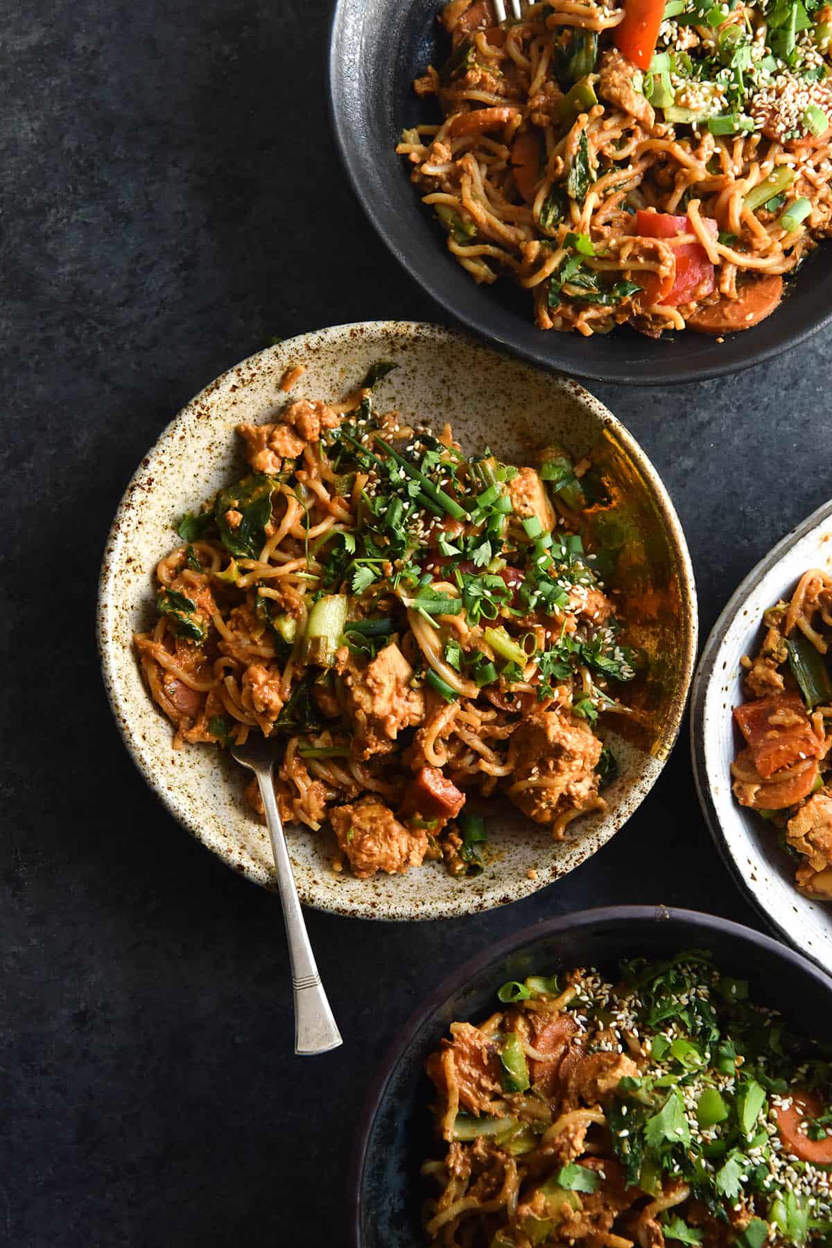 An aerial view of four ceramic bowls filled with FODMAP friendly peanut butter stir fry. The stir fry is filled with chunks of tofu and vegetables and topped with sesame seeds and spring onions. The bowls are all different colours and shapes, sitting atop a dark blue backdrop and aligned to the right of the image