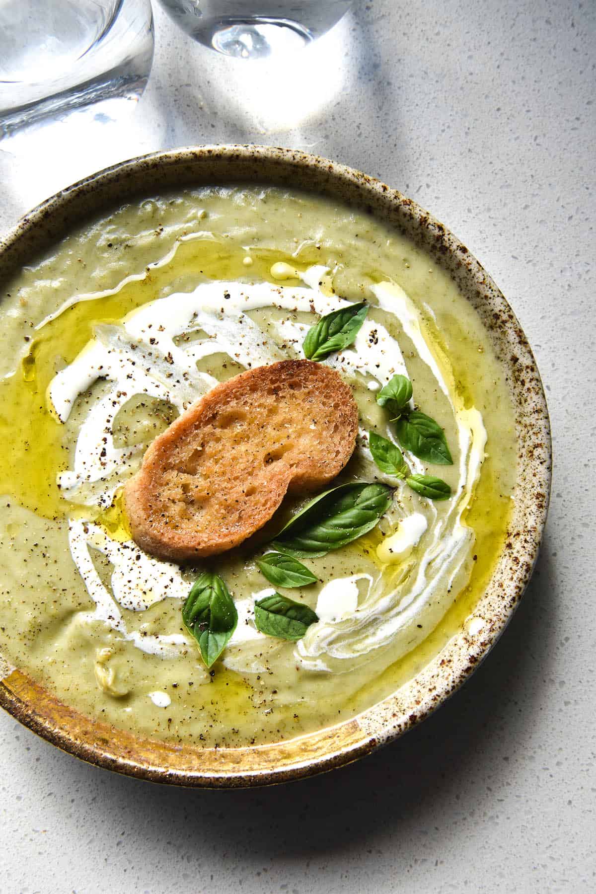 An aerial view of a speckled ceramic bowl of FODMAP friendly leek and potato soup. The soup is a light green because it uses leek greens to keep it FODMAP friendly. It is topped with a swirl of cream, truffle oil, a gluten free crouton, grilled haloumi and basil leaves.