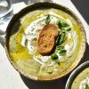 An aerial view of a speckled ceramic bowl of FODMAP friendly leek and potato soup. The soup is a light green because it uses leek greens to keep it FODMAP friendly. It is topped with a swirl of cream, truffle oil, a gluten free crouton, grilled haloumi and basil leaves. The bowl sits in harsh sunlight on a white table. Two water glasses sit in the top right corner of the image, while another bowl of soup sits in the bottom lefthand corner.