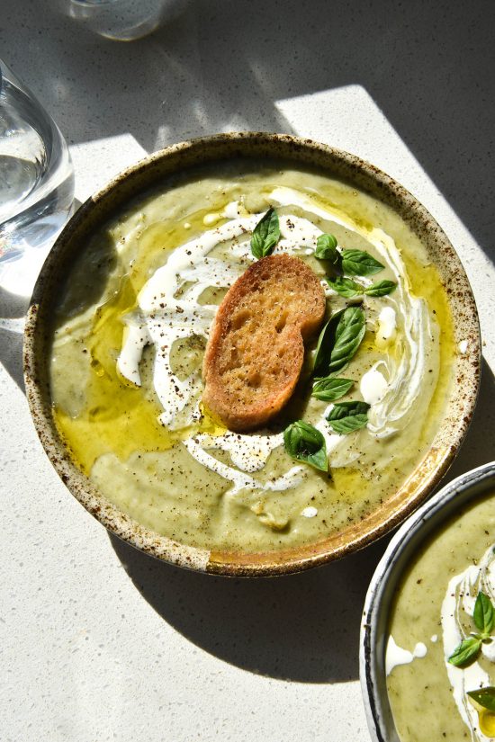 An aerial view of a speckled ceramic bowl of FODMAP friendly leek and potato soup. The soup is a light green because it uses leek greens to keep it FODMAP friendly. It is topped with a swirl of cream, truffle oil, a gluten free crouton, grilled haloumi and basil leaves. The bowl sits in harsh sunlight on a white table. Two water glasses sit in the top right corner of the image, while another bowl of soup sits in the bottom lefthand corner.
