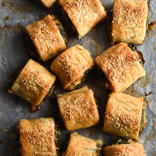 An aerial view of a tray of gluten free spinach and feta rolls. The rolls are casually strewn in a bundle on a baking paper lined baking tray. They are golden brown and topped with toasted sesame seeds