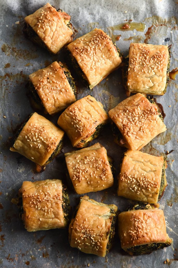 An aerial view of a tray of gluten free spinach and feta rolls. The rolls are casually strewn in a bundle on a baking paper lined baking tray. They are golden brown and topped with toasted sesame seeds