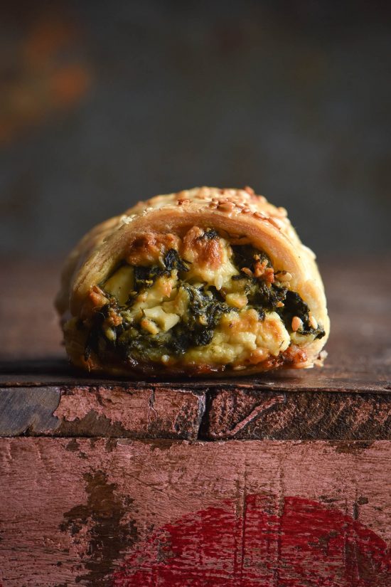 A side on macro photo of a gluten free spinach and feta sausage roll. The roll sits atop a pink and red vintage wooden box and in front of a mottled rusting background. Golden feta and pine nuts poke out of the filling, and the top of the pastry is sprinkled with toasted sesame seeds.